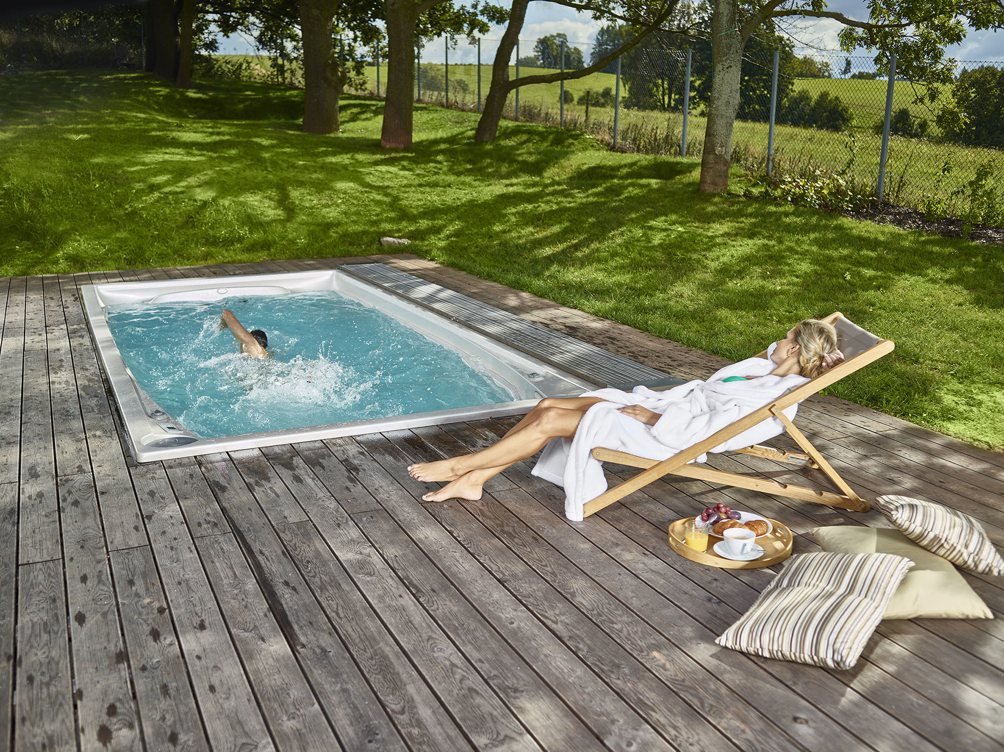 Frau in Bademantel entspannt sich auf einer Holzterrasse neben einem hochwertigen USSPA Swim Spa, während jemand darin schwimmt – idyllische Outdoor-Spa-Umgebung für Wellness und Entspannung.
